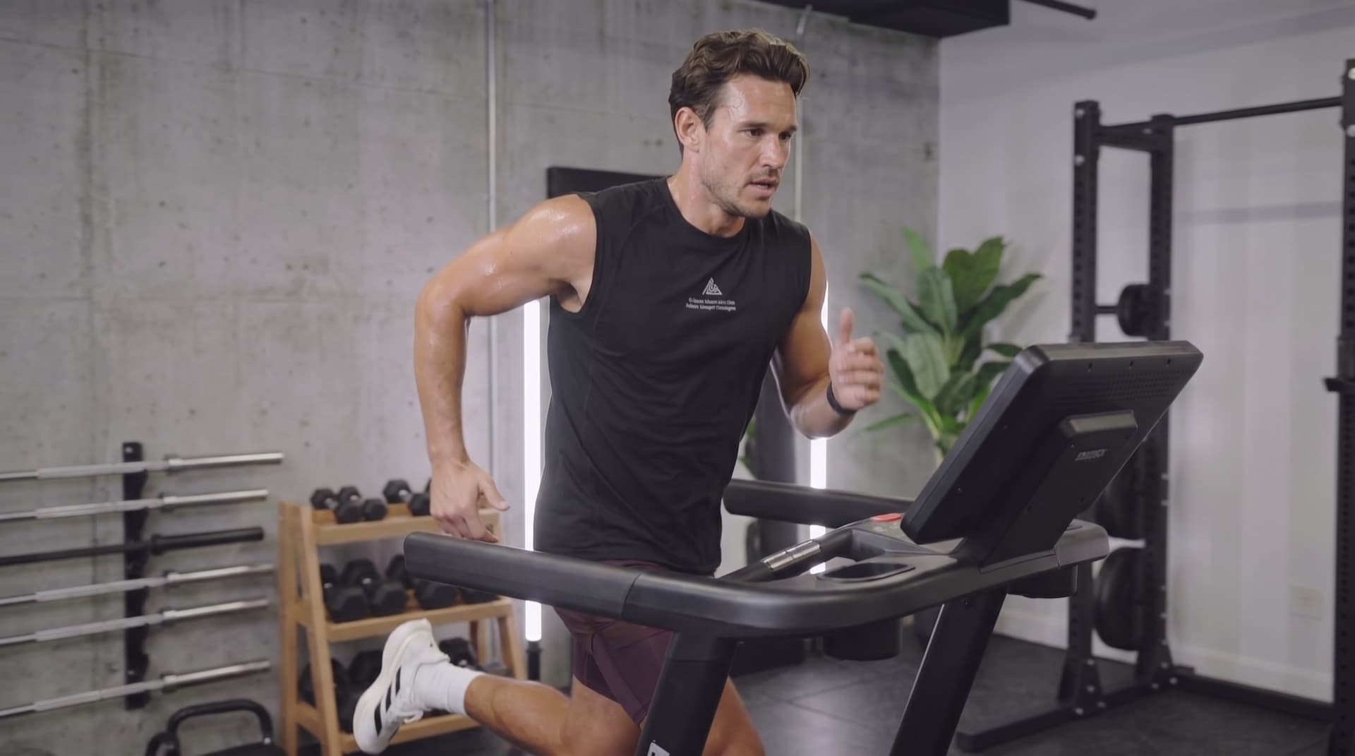 Hybrid athlete mid-stride on a treadmill in a concrete-walled gym, dumbbells and a power rack in the background